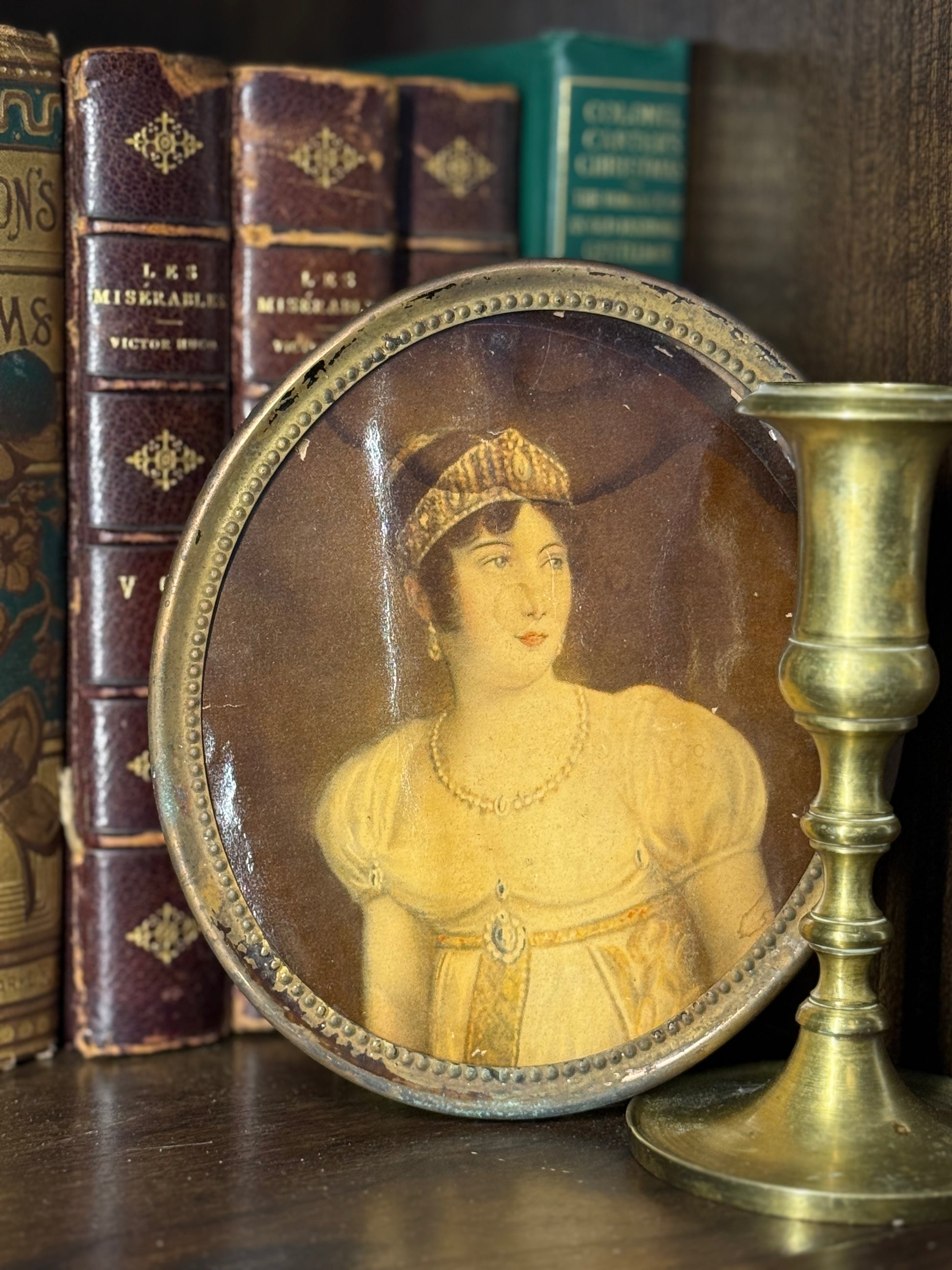 Small round portrait of a woman in a decorative frame on a wooden surface with books and a candlestick.
